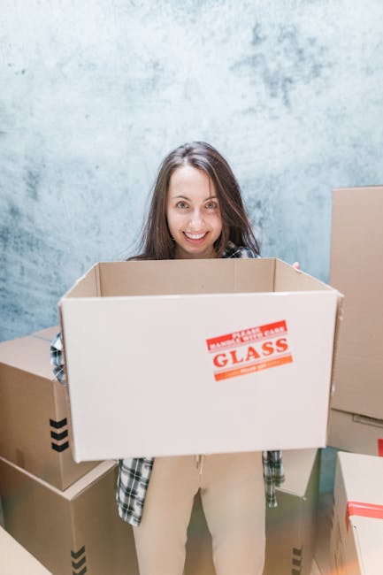 A woman with long brown hair smiling indoors, holding an open cardboard moving box labeled 'GLASS' in red and white, during a home relocation process. She is standing in front of a plain, light blue textured wall, surrounded by several other cardboard boxes of various sizes, some sealed with packing tape, indicating packing activities associated with house removals. The boxes are made of standard brown corrugated cardboard, with some wrapped in plastic film for protection. The scene appears to take place in a residential setting, with natural or soft ambient lighting illuminating the space. The presence of multiple packed boxes suggests the early stages of furniture transport and packing for a professional move, aligning with the services offered by Man with Van Mayfair for residential moves in the Mayfair area.