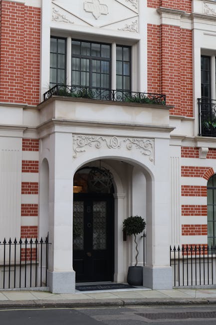 The image depicts the exterior of a stately residential building with a red brick and white stone facade, featuring intricate decorative detailing above the arched entranceway. The entrance has a black door with a frosted glass panel, flanked by two potted plants with neatly trimmed foliage placed in dark containers. Above the doorway, there is a balcony with a black wrought iron railing and a window with black framing. The scene shows a sidewalk with a metal fence along the building's perimeter, and the overall setting suggests a high-end urban context typical of a London district such as Mayfair. The architectural style and arrangement of elements imply that this location may be associated with professional house removal or moving services, such as those provided by Man with Van Mayfair, including moving preparations or logistical arrangements for home relocation.