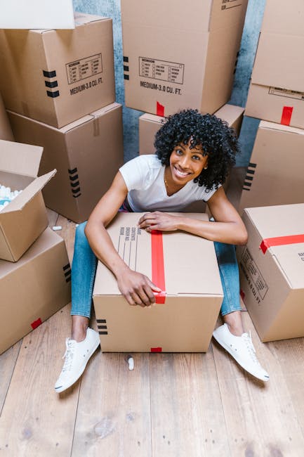 A woman with long brown hair smiling indoors, holding an open cardboard moving box labeled 'GLASS' in red and white, during a home relocation process. She is standing in front of a plain, light blue textured wall, surrounded by several other cardboard boxes of various sizes, some sealed with packing tape, indicating packing activities associated with house removals. The boxes are made of standard brown corrugated cardboard, with some wrapped in plastic film for protection. The scene appears to take place in a residential setting, with natural or soft ambient lighting illuminating the space. The presence of multiple packed boxes suggests the early stages of furniture transport and packing for a professional move, aligning with the services offered by Man with Van Mayfair for residential moves in the Mayfair area.
