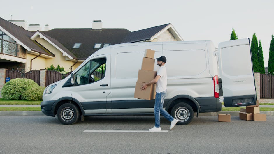 A man wearing a white t-shirt, jeans, white sneakers, and a black cap is unloading cardboard boxes from a white cargo van parked on a residential street. The van is positioned parallel to the pavement
