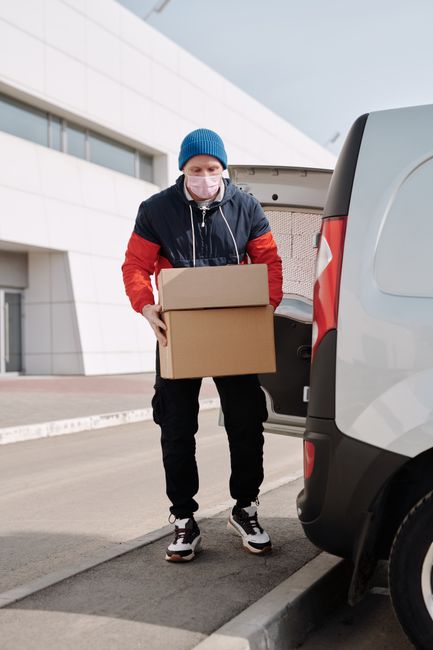 A man wearing a white t-shirt, jeans, white sneakers, and a black cap is unloading cardboard boxes from a white cargo van parked on a residential street. The van is positioned parallel to the pavement