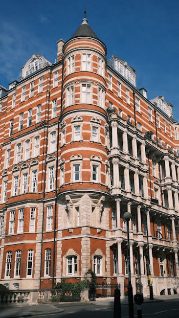 Exterior view of a historic, multi-storey building with ornate white stone detailing and red brickwork, situated on a city street under a clear blue sky. In the foreground, a resident or mover's trolley loaded with several large cardboard boxes, some wrapped in plastic and fabric blankets for protection, is positioned near the doorway of the building. A person dressed in casual clothing is seen lifting or carrying a light wooden piece of furniture, such as a small table or cabinet, away from the entrance, indicating a home relocation process. Nearby, a vehicle, likely a van used by [COMPANY_NAME], is parked on the paved street close to the building, ready for the furniture transport part of the moving service. The scene captures a typical moving day with objects prepared for packing and loading, illustrating the logistics involved in residential removals or furniture transport in a well-preserved Mayfair property.