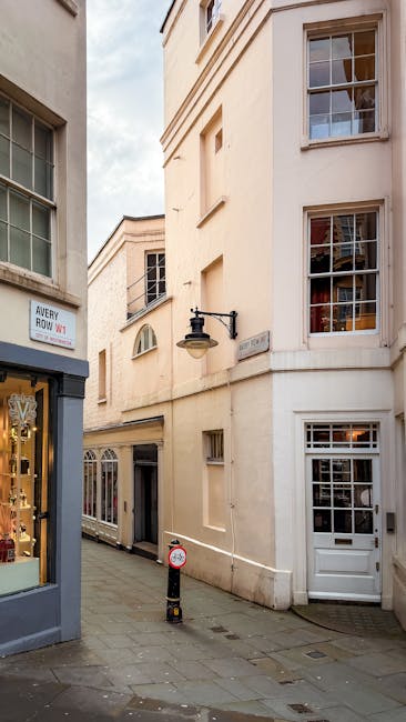 A multi-storey brick building with black wrought iron balconies and white-framed windows on a Mayfair street. Two large Union Jack flags are mounted on ropes above the entrance, which has a black canopy with the sign 'Claridge's' visible. In front of the building, there is a black metal fence with decorative elements, and pink flowering plants along the pavement. On the street, a black taxi and a blue vehicle are parked, while a person is seen walking nearby. A traffic light is positioned at the corner of the street, and the scene suggests an urban environment suitable for house removals or furniture transport, potentially involving a home relocation or moving services provided by Man with Van Mayfair.