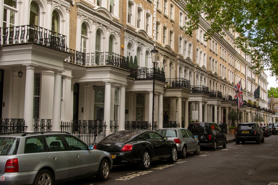 A row of elegant white Victorian-style townhouses with decorative black wrought iron balconies and columns, situated on a tree-lined street in Mayfair. Several cars, including a silver estate car, a black sports car, and other vehicles, are parked along the pavement in front of the properties. The image shows a typical urban residential setting with a lamppost and potted plants on some balconies, as well as Union Jack flags hanging from one of the buildings. This scene illustrates a typical house exterior during daytime, with natural light and lush greenery in the foreground, representing a setting suitable for home relocation or furniture transport services by Man with Van Mayfair, as part of their removals and packing logistics.