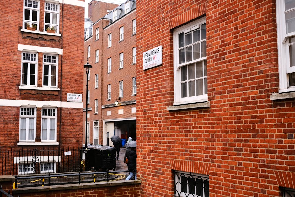 The image shows the exterior of a red brick building in an urban setting, with three visible white-framed sash windows on the upper floors. A street sign reading 'PROVIDENCE COURT W1' is mounted on the building wall, along with another smaller sign indicating 'BALDERSTON STREET W1.' In the foreground, a black metal fence surrounds a small lower-level window, and a flashing yellow and black tape marks a restricted area near the pavement. Two individuals, dressed in casual clothing and wearing hooded jackets, are seen carrying boxes and large plastic bags, suggesting a home relocation or moving process. Behind them, a black moving van with open rear doors is parked against a narrow alleyway, with packing materials and boxes visible near the vehicle, indicating loading or unloading activities. The scene is lit with natural daylight, highlighting the typical London residential environment associated with professional removals services such as those provided by Man with Van Mayfair.