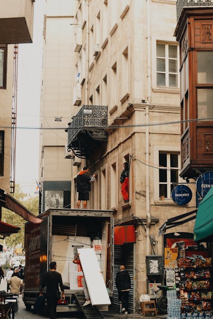 The image shows a narrow urban street scene outside a multi-storey building with beige stone facade, featuring several small rectangular windows and a wrought iron balcony partially visible on the upper floor. A man dressed in dark clothing with an orange safety helmet is on a scaffold platform attached to the building, handling a large piece of fabric or wrapping material, which may be used for packing furniture during a home relocation. Below, an open moving van with its rear doors down is parked on the street, containing visible packing supplies such as cardboard boxes and protective blankets. Several people are present: one standing near the van, possibly overseeing the loading process, and others walking along the pavement. To the right, a building with a wooden exterior and a shop storefront displays various items, including snacks and bottled water, under a green awning. A clothesline with textiles or clothing hangs across the street, emphasizing a busy, lived-in environment typical of a city with active move-related activities. The scene is captured during daylight, with natural light illuminating the surroundings, showcasing a typical move day involving furniture transport and packing close to the building’s loading area. Man with Van Mayfair is operating within this setting, assisting with the logistics of home relocation and furniture transport in the busy urban environment.