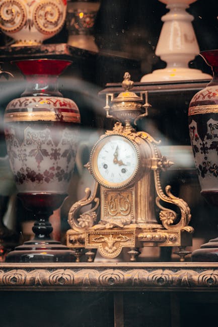 A close-up view of an ornate antique clock with a white dial and decorative gold detailing, positioned on a wooden surface among several large ceramic vases featuring intricate floral patterns in red, black, and white. The clock is surrounded by other decorative objects, including a white ceramic lamp and additional vases in a setting that appears to be inside a shop or storage area for household antiques. The image captures the detailed craftsmanship of the clock and vases, with some reflections and soft lighting highlighting the textures and finishes. This scene relates to the process of home relocation or furniture transport, as [COMPANY_NAME], such as Man with Van Mayfair, often handles the careful packing and moving of valuable and delicate items like antiques during house removals or relocation services in Mayfair.