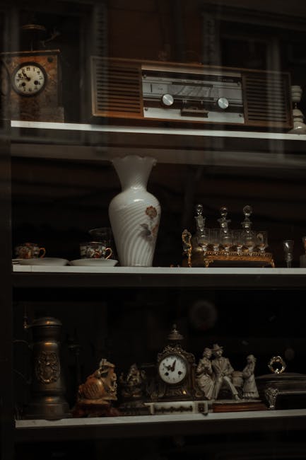 A close-up view of a glass-fronted wooden cabinet inside a house during a furniture removal process. The cabinet contains various decorative items, including an antique-style clock, ceramic figurines, a vintage radio placed on a higher shelf, and ornate glass decanters with stoppers. The shelving is filled with fine crockery, small sculptures, and metallic ornaments, all arranged neatly. The scene is lit with soft indoor lighting, highlighting the details of the decorative objects. This setting reflects part of a home interior where detailed packing and safeguarding of delicate antique furnishings are essential for a house relocation. Man with Van Mayfair, a specialist in removals and furniture transport, is overseeing the careful handling of these items as part of a professional moving service, ensuring safe packing, loading, and transport of valuable, bulky antiques during the home move.