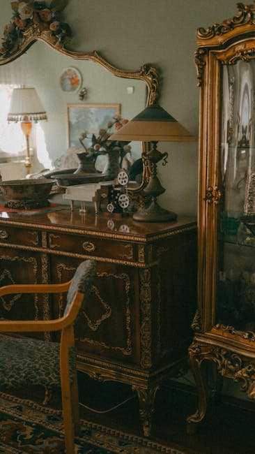A close-up view of an ornate antique clock with a white dial and decorative gold detailing, positioned on a wooden surface among several large ceramic vases featuring intricate floral patterns in red, black, and white. The clock is surrounded by other decorative objects, including a white ceramic lamp and additional vases in a setting that appears to be inside a shop or storage area for household antiques. The image captures the detailed craftsmanship of the clock and vases, with some reflections and soft lighting highlighting the textures and finishes. This scene relates to the process of home relocation or furniture transport, as [COMPANY_NAME], such as Man with Van Mayfair, often handles the careful packing and moving of valuable and delicate items like antiques during house removals or relocation services in Mayfair.