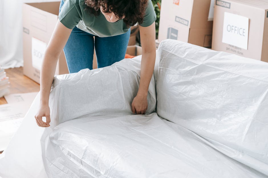 A person wearing a green top and blue jeans is inside a room, wrapping a piece of furniture with protective white plastic sheeting for a house move, with several cardboard boxes and packing materials visible in the background. The furniture appears to be a sofa or armchair, with the plastic covering being carefully fitted around its shape. The setting suggests a packing process as part of a home relocation, assisted by Man with Van Mayfair, with professional handling of fragile or bulky items to ensure safe transportation. The room is well-lit and organized, with some packing boxes labeled for different rooms, and the individual is focused on securely wrapping the furniture to prevent damage during the logistics of furniture transport and removal services.