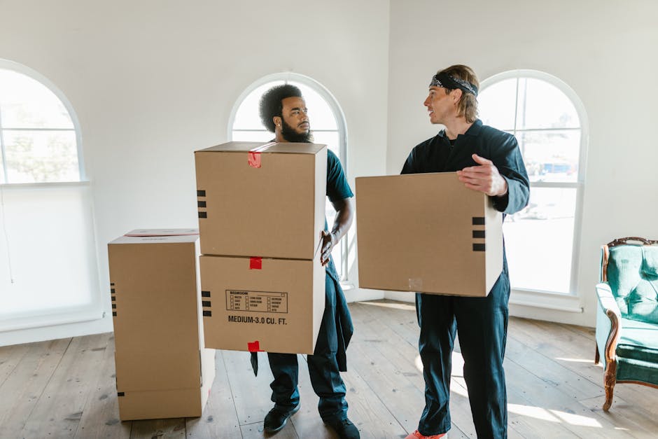A young woman sitting cross-legged on a wooden floor inside a room during a home relocation process, surrounded by multiple large cardboard boxes sealed with tape and some labeled with fragile stickers. She is wearing casual clothing including a white tank top and sneakers, and is smiling while tossing packing peanuts into the air. The boxes are stacked in various positions, with some on the floor and others against the wall, which has a textured light blue finish. In the background, a small shelf with decorative items and a potted plant are visible, along with a doorway leading to another room. The scene depicts the packing and moving stage typical of furniture transport and home removals, with [COMPANY_NAME] facilitating the logistics of transition within the Mayfair area as described in the page titled 'W1K Removals: Best Practices for Mayfair Postcode Moves'.